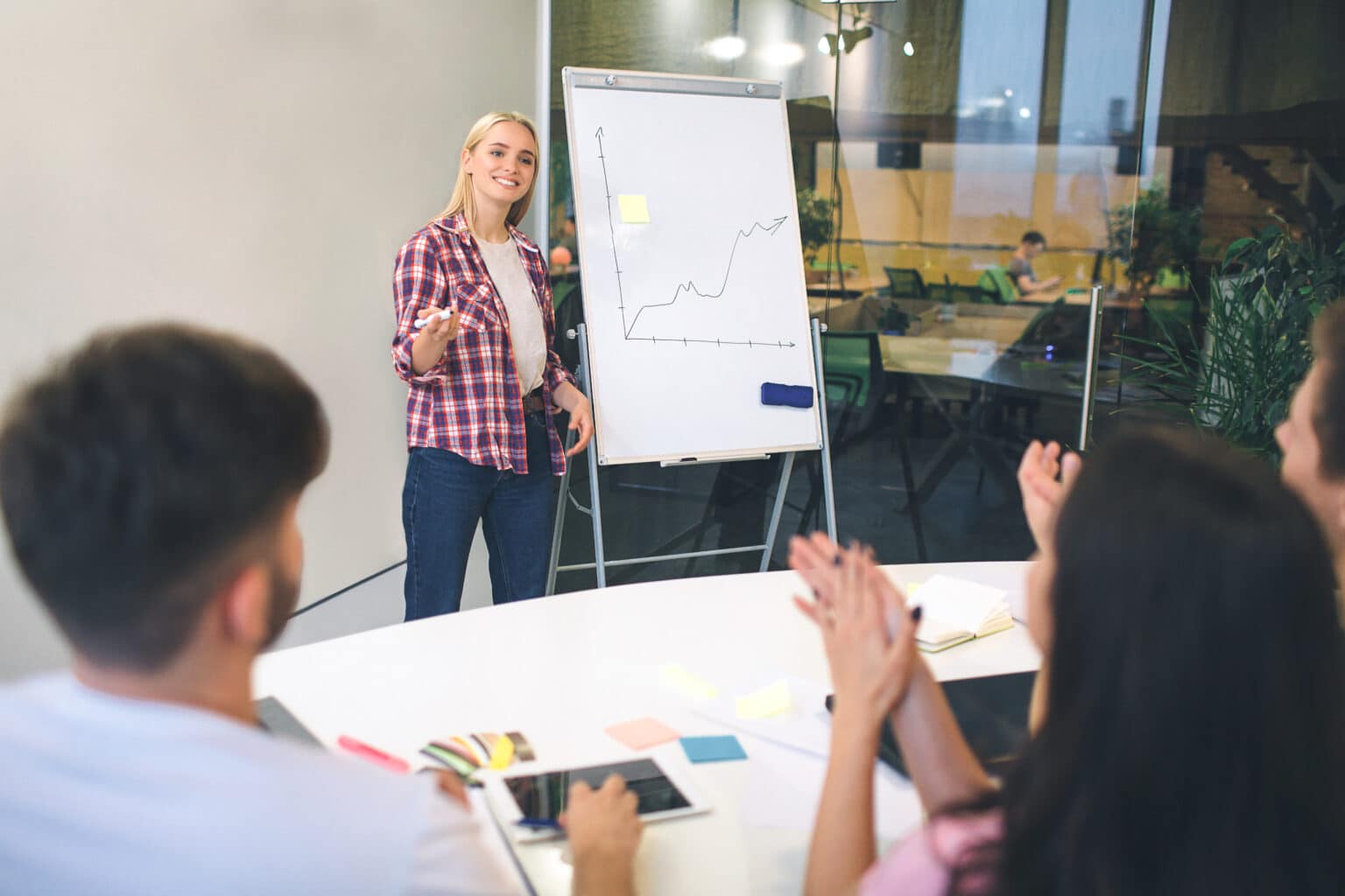 Cheerful young blonde woman stand at flipchartand smile. She point at people who sit and have conversation. They all are in room